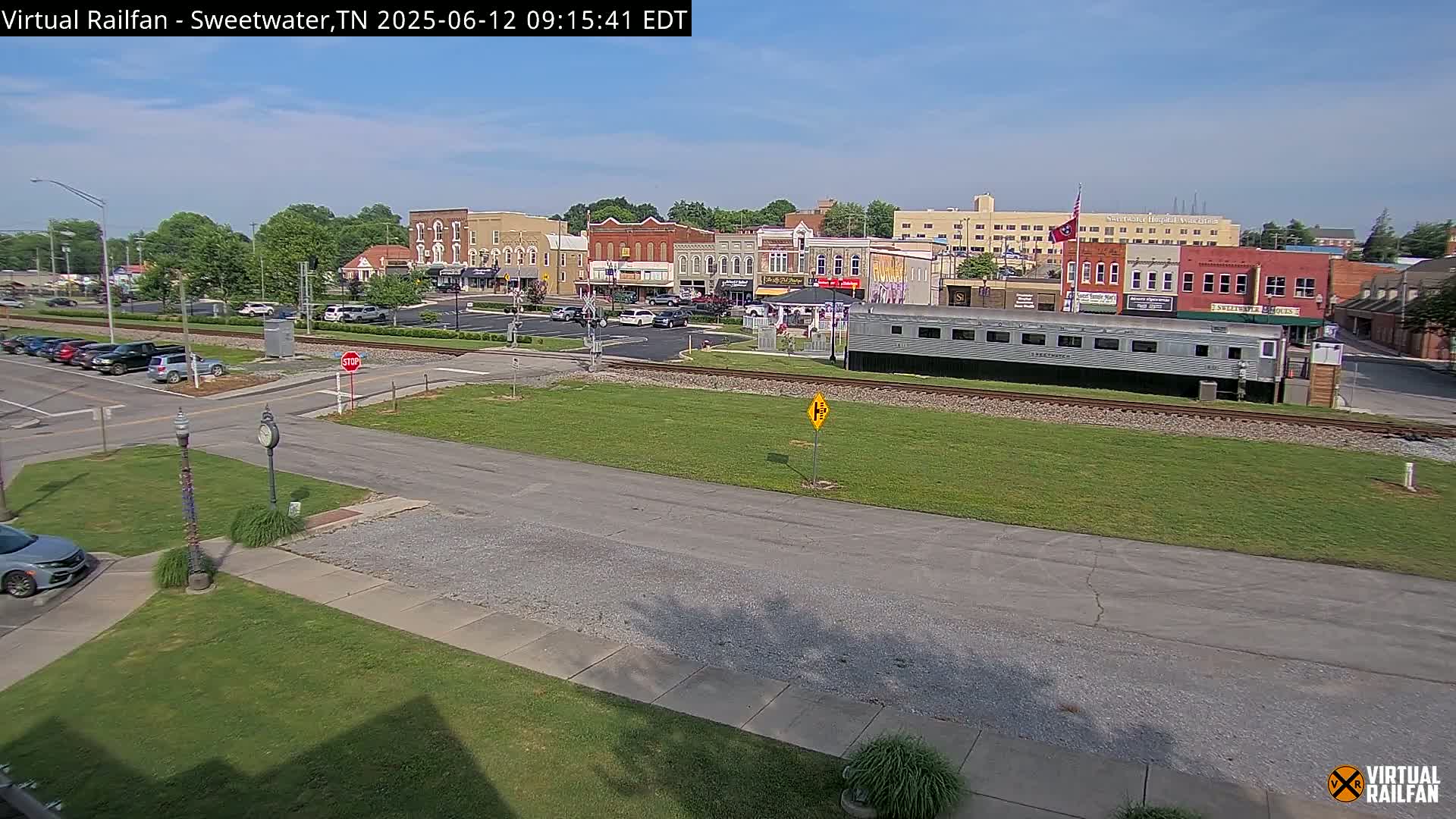 A mostly sunny day reveals a small town's downtown area, featuring several brick buildings, a parking lot, and a long silver train car sitting on train tracks in the foreground.