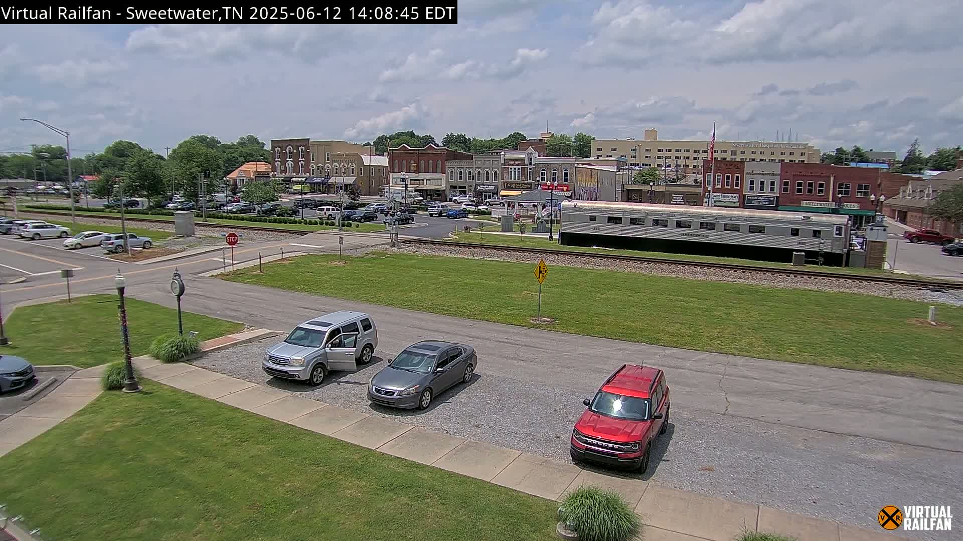 A partly sunny day shows a small town square with several cars parked, a silver passenger train car, and several brick buildings.
