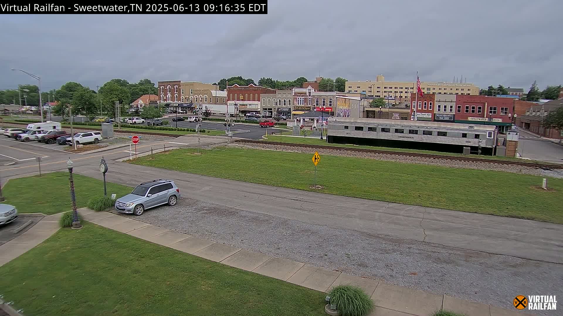 A mostly overcast day shows a small town's square with several brick buildings, a parked silver SUV, and a long silver train car sitting on train tracks in the foreground.