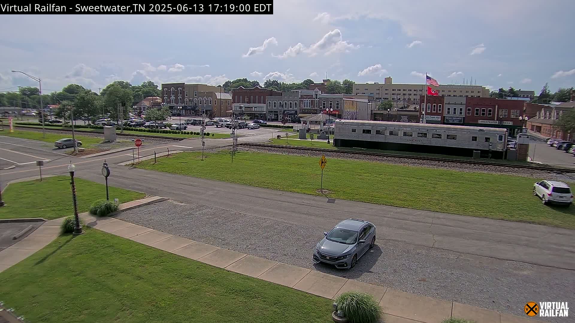 A mostly sunny view of a town square with parked cars, a railroad track, and a silver passenger rail car, next to several brick buildings.