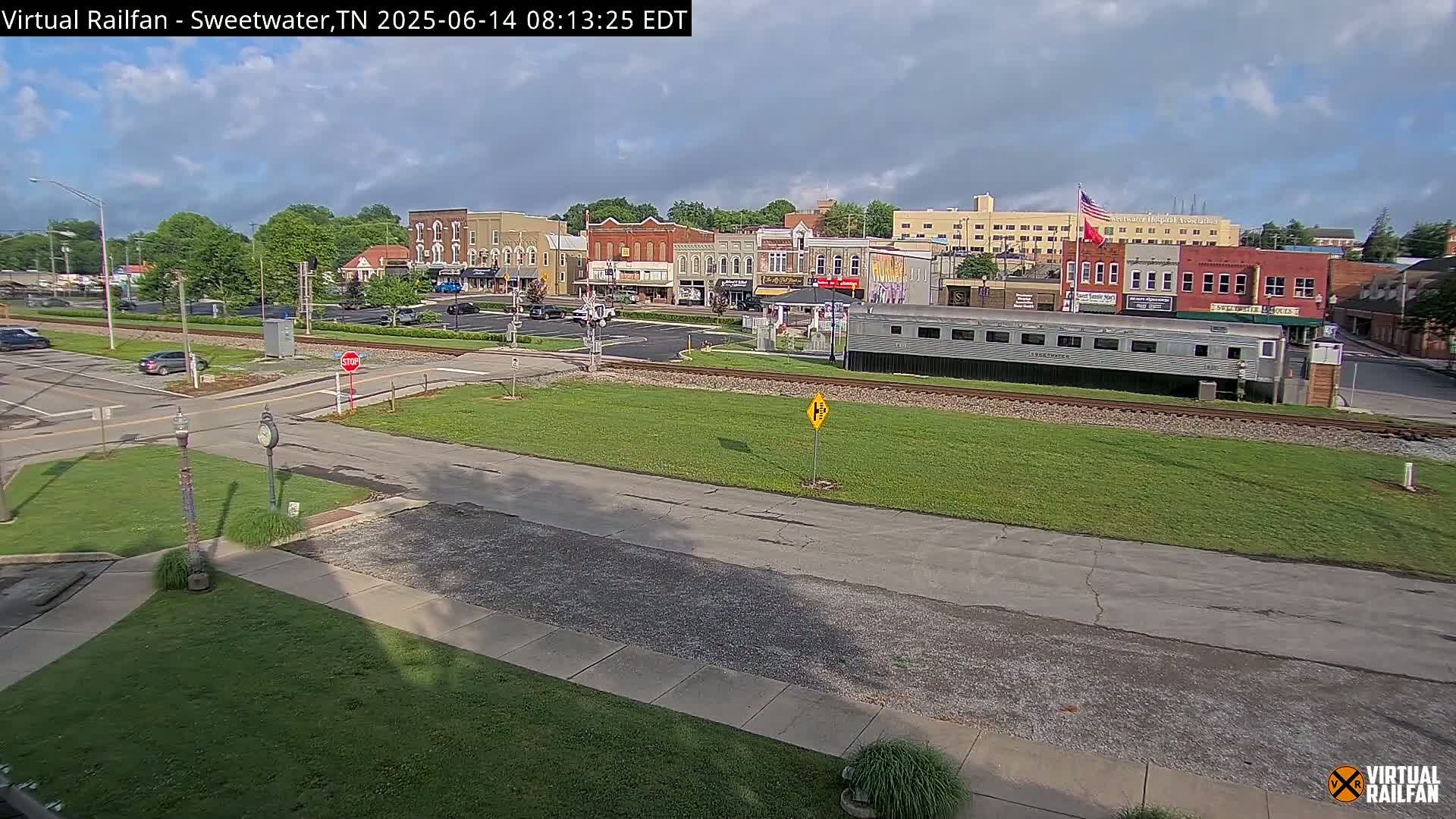 A partly cloudy day reveals a small town square with several brick buildings, a railway track with a long silver train car, and a grassy area.
