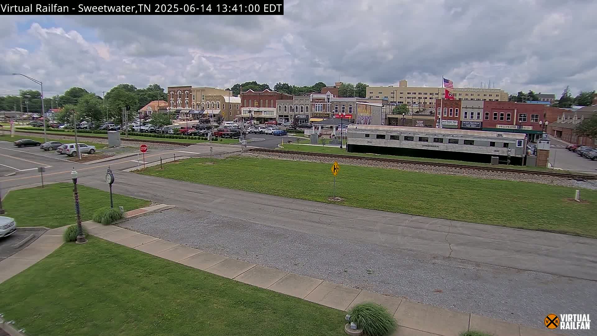 A partly cloudy day shows a small town's downtown area with parked cars, a railroad crossing, and a long silver train car sitting on the tracks.