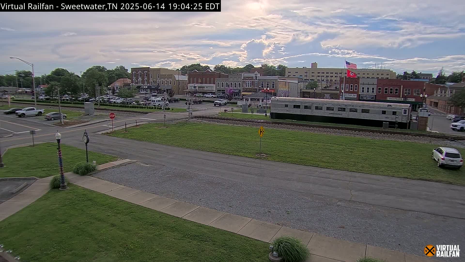 A partly cloudy day shows a small town's downtown area with parked cars, a railroad crossing, and a long silver train car sitting on the tracks.