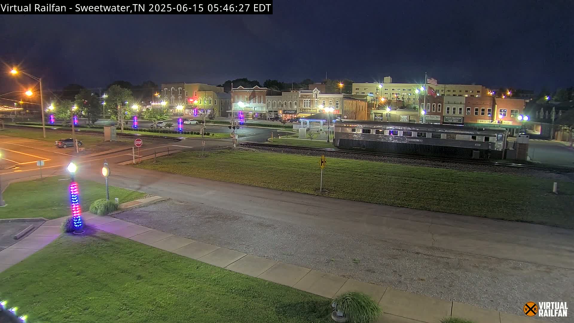 A nighttime view of a small town's downtown area, with a train car parked on tracks in the foreground, and decorative light poles lining a street.