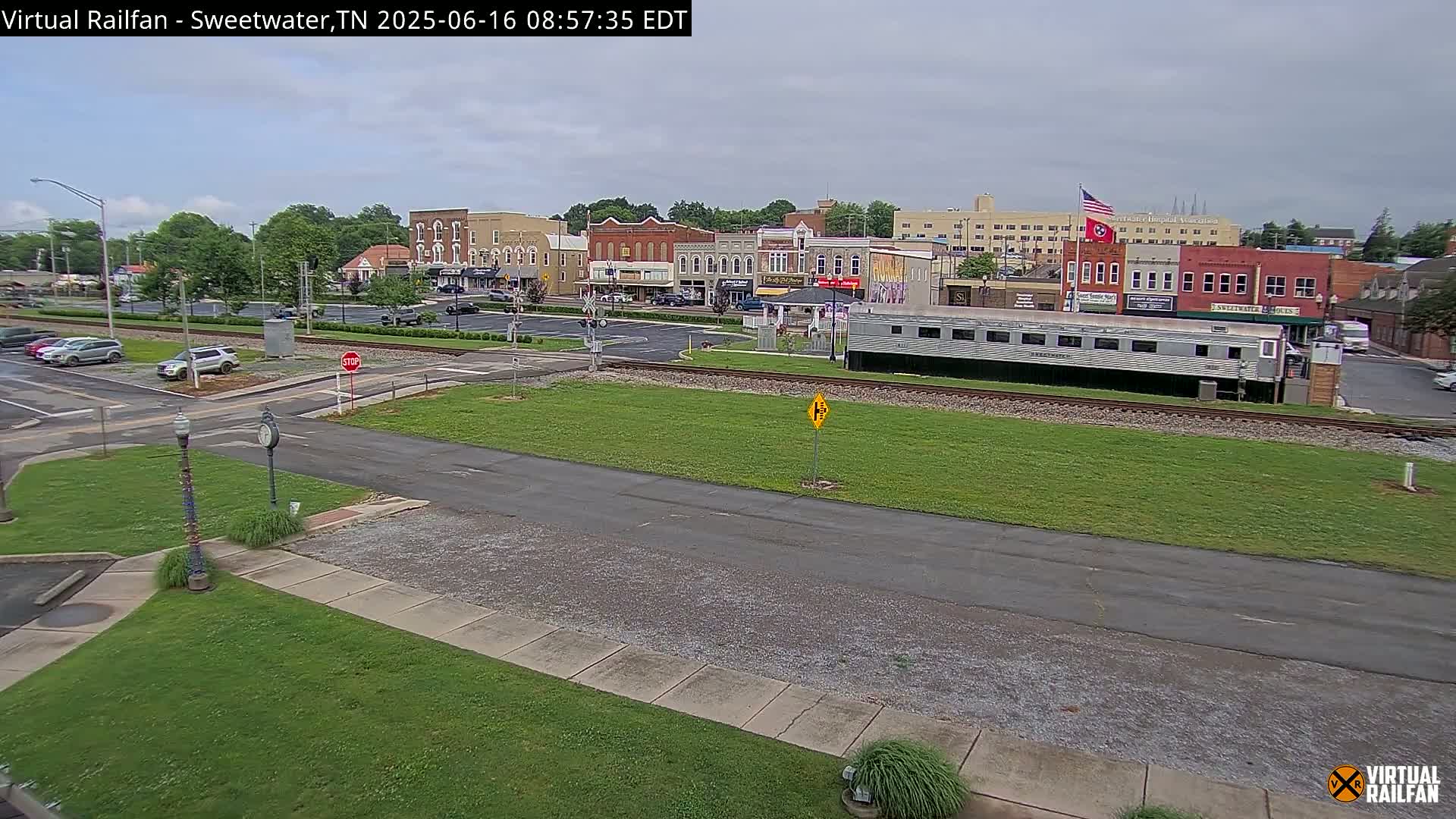A small town's downtown area, under an overcast sky, features several brick buildings, a railway track with a passenger car parked beside it, and sparse car traffic.