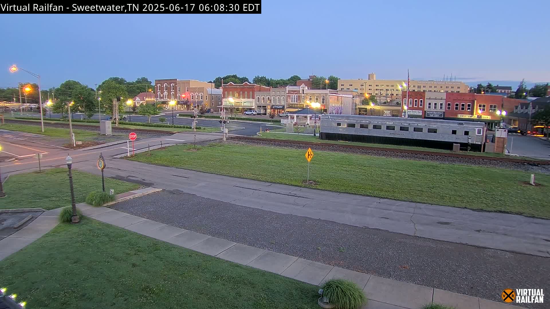 A twilight view of a small town's downtown area, featuring a silver passenger railcar parked near the train tracks,  with  grassy areas and roads surrounding the buildings.