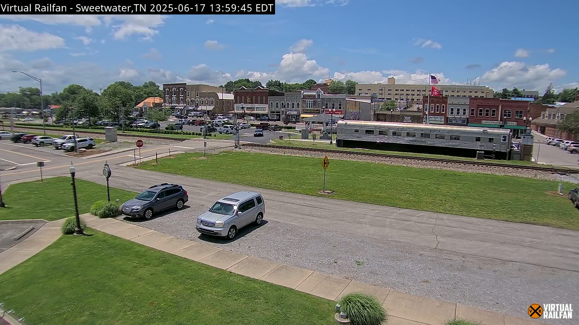 A sunny day reveals a small town square with several cars parked near a railway track where a long passenger car sits, with a background of brick buildings.