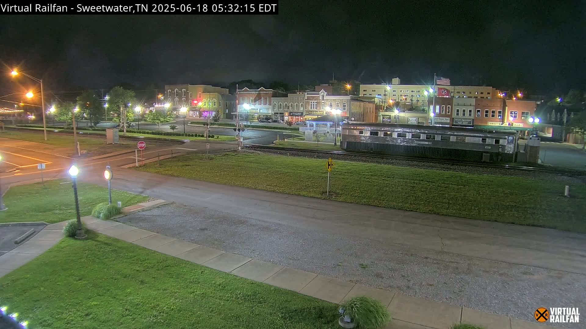A nighttime view of a town square with several buildings, a railway track with a train car, and a grassy area.