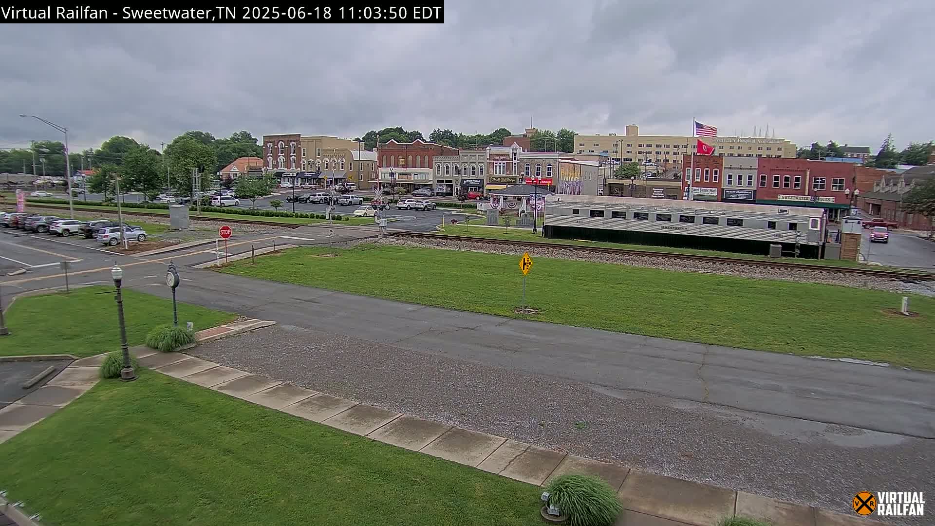 A mostly cloudy day reveals a small town's downtown area, featuring several brick buildings, a railway line with a parked train car, and a street with some cars.