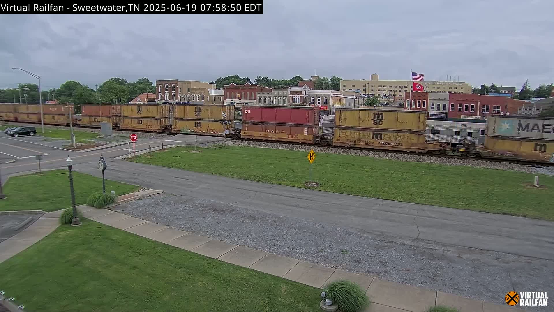 A long freight train sits on the tracks near a road intersection in front of a town under an overcast sky.