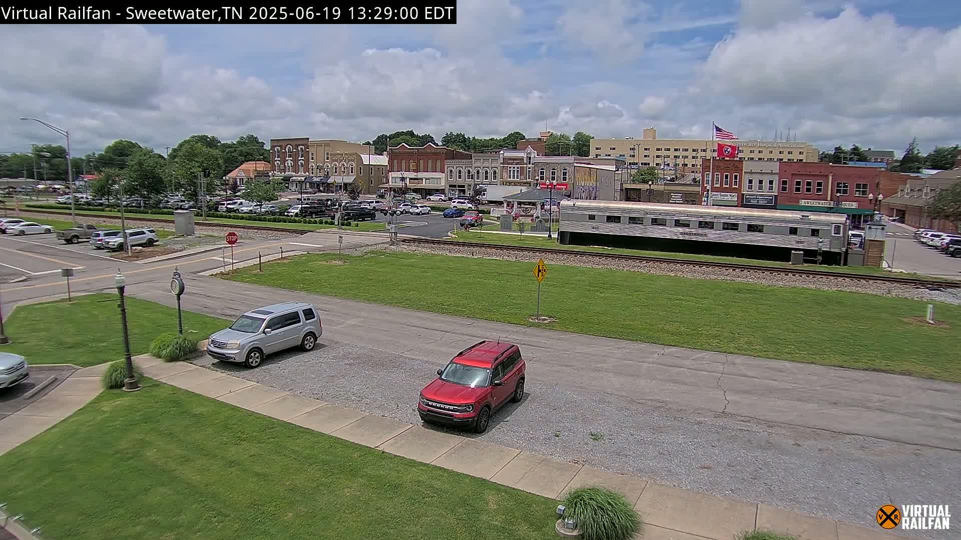 A partly cloudy day reveals a small town square with parked cars, a silver railway car, and a few vehicles in a parking lot, all situated near a railroad track.