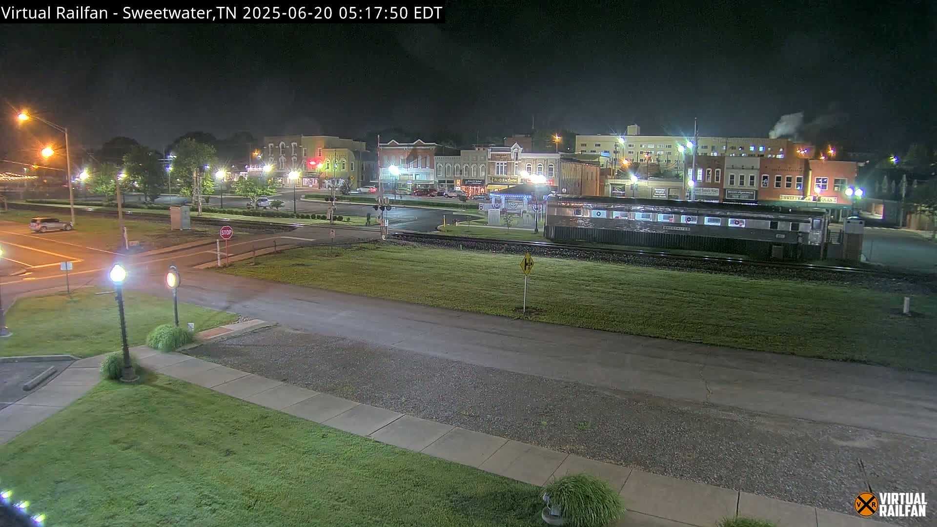 A nighttime view of a small town's downtown area, featuring several buildings, a railway crossing, a train car, and sparsely traveled streets under clear skies.