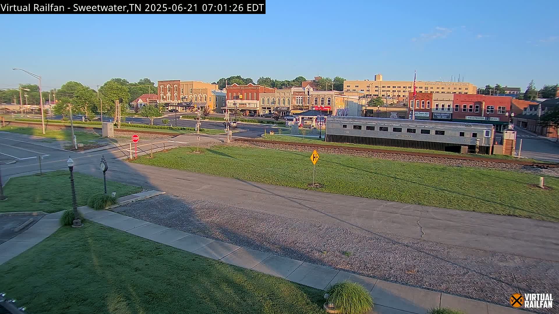 A sunny morning view of a small town's downtown area, featuring several brick buildings, a railway track with a long silver train car parked beside it, and a grassy area in the foreground.