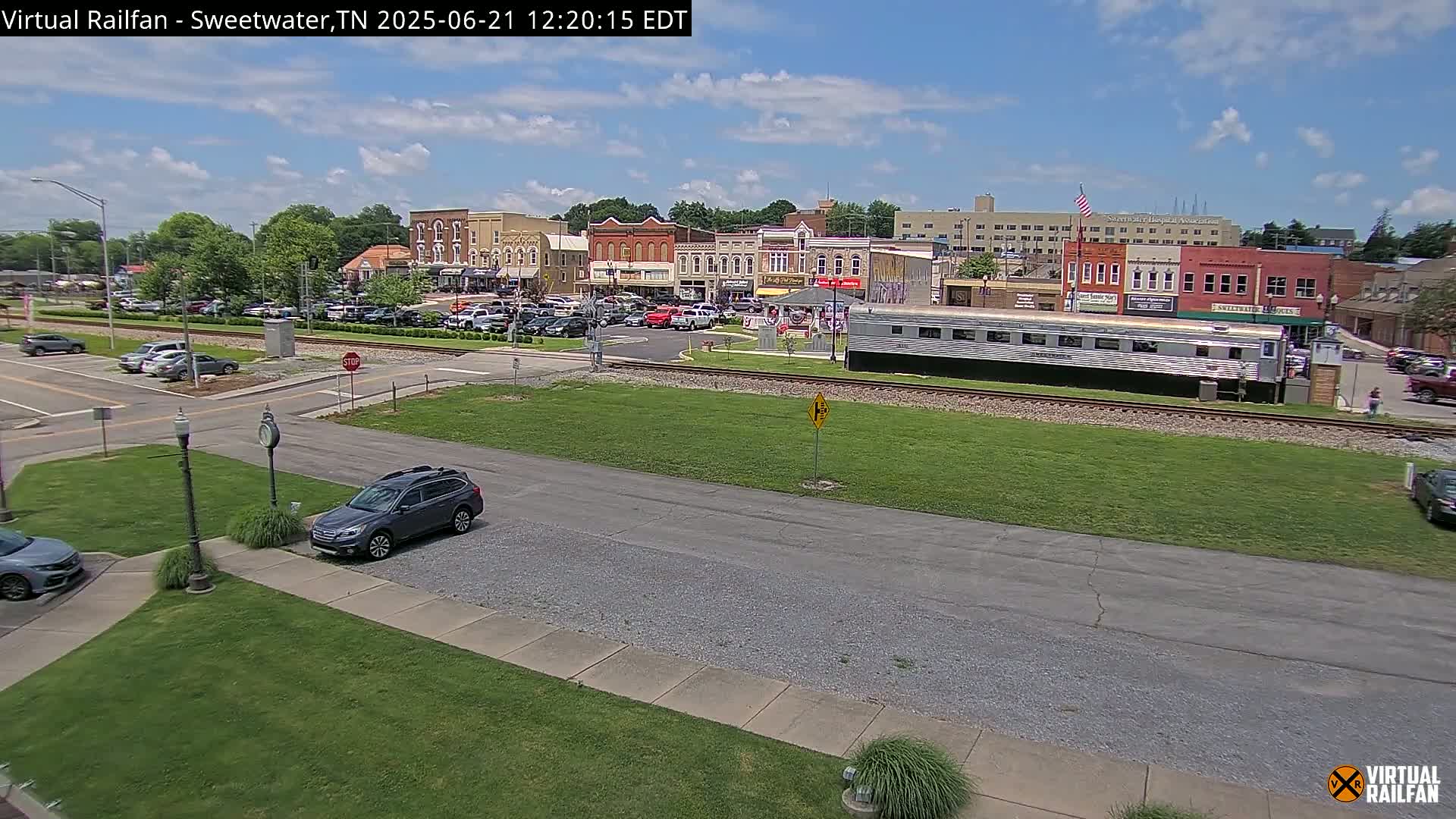 A sunny day reveals a small town's downtown area, with a train car parked on tracks in front of brick buildings and cars parked in lots.