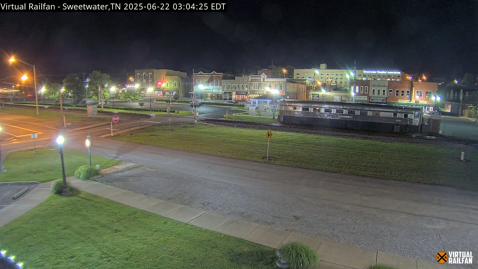 A nighttime view of a small town's downtown area, featuring a train car parked on a railway track near a street intersection and several buildings, all lit under dark skies.