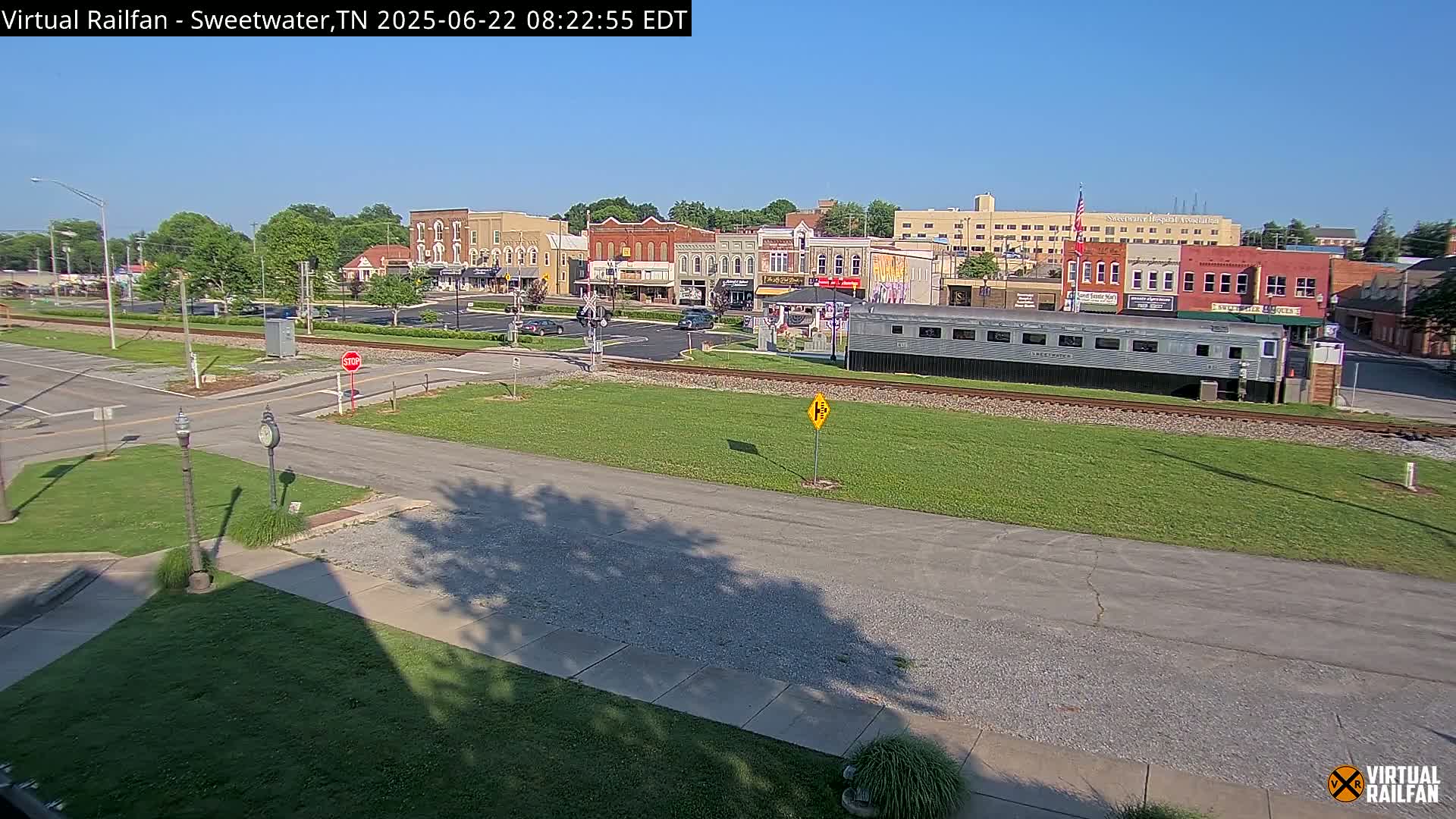 A sunny view of a small town's downtown area, featuring several brick buildings, a railroad crossing, and a long silver train car parked on the tracks.