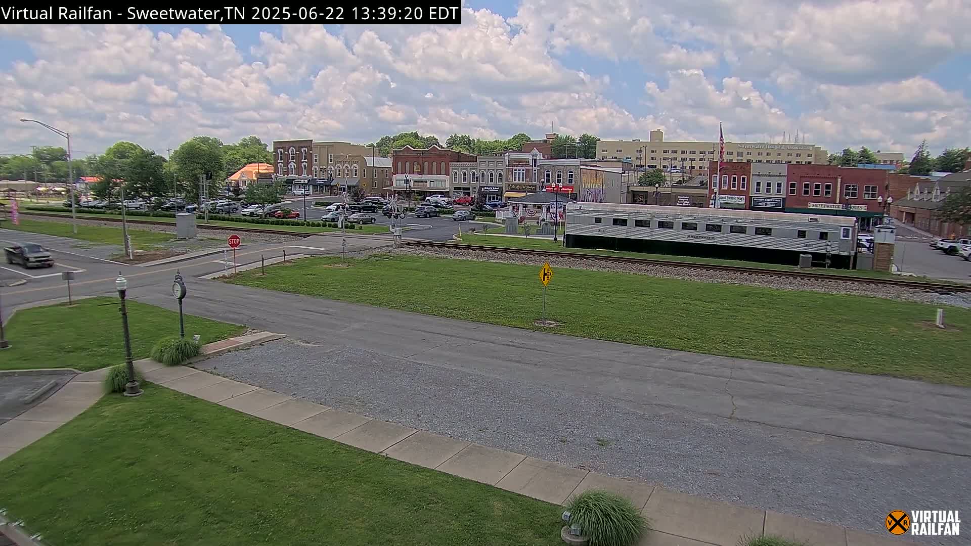 A partly cloudy day shows a small town's downtown area with several buildings, cars parked along the street, and a long train car sitting on the railroad tracks.