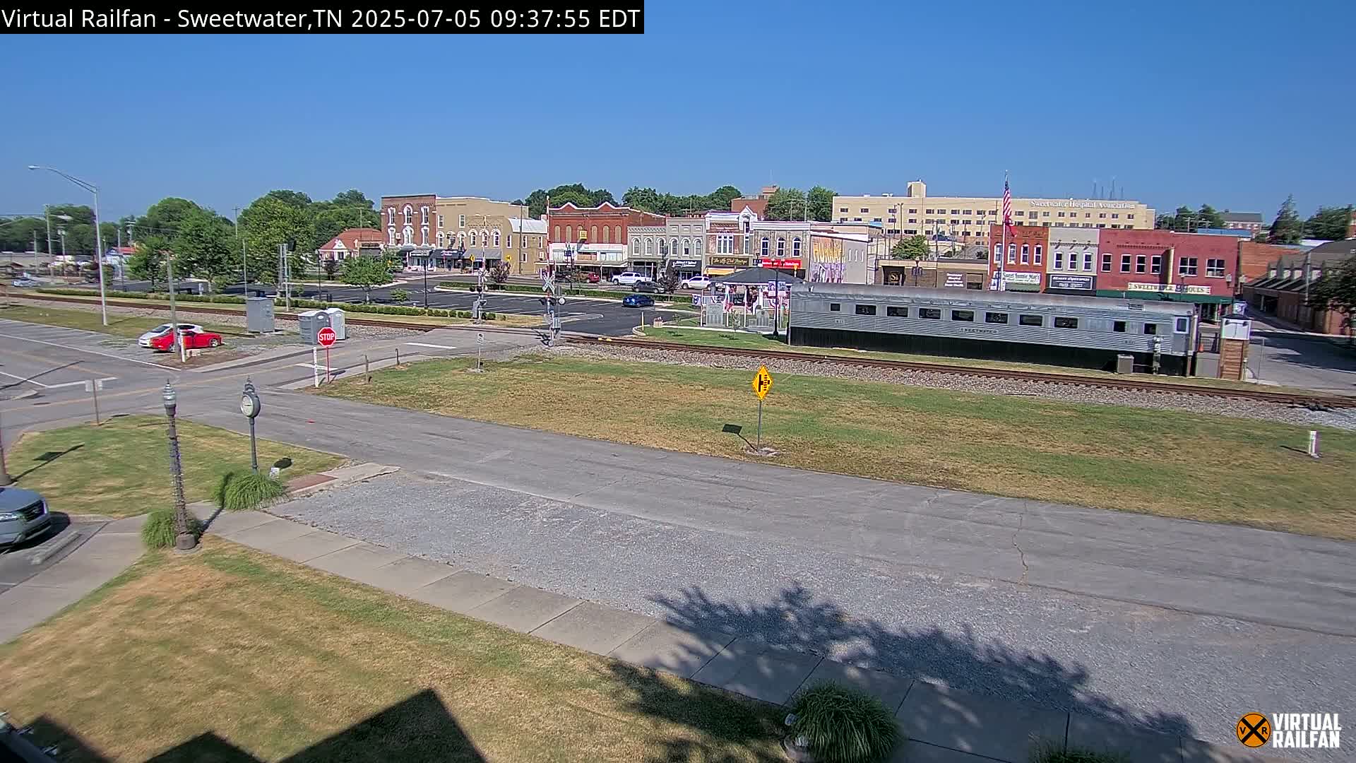 A mostly sunny day reveals a small town's downtown area, featuring brick buildings, a railroad crossing with a parked train car, and a few cars in a parking lot.