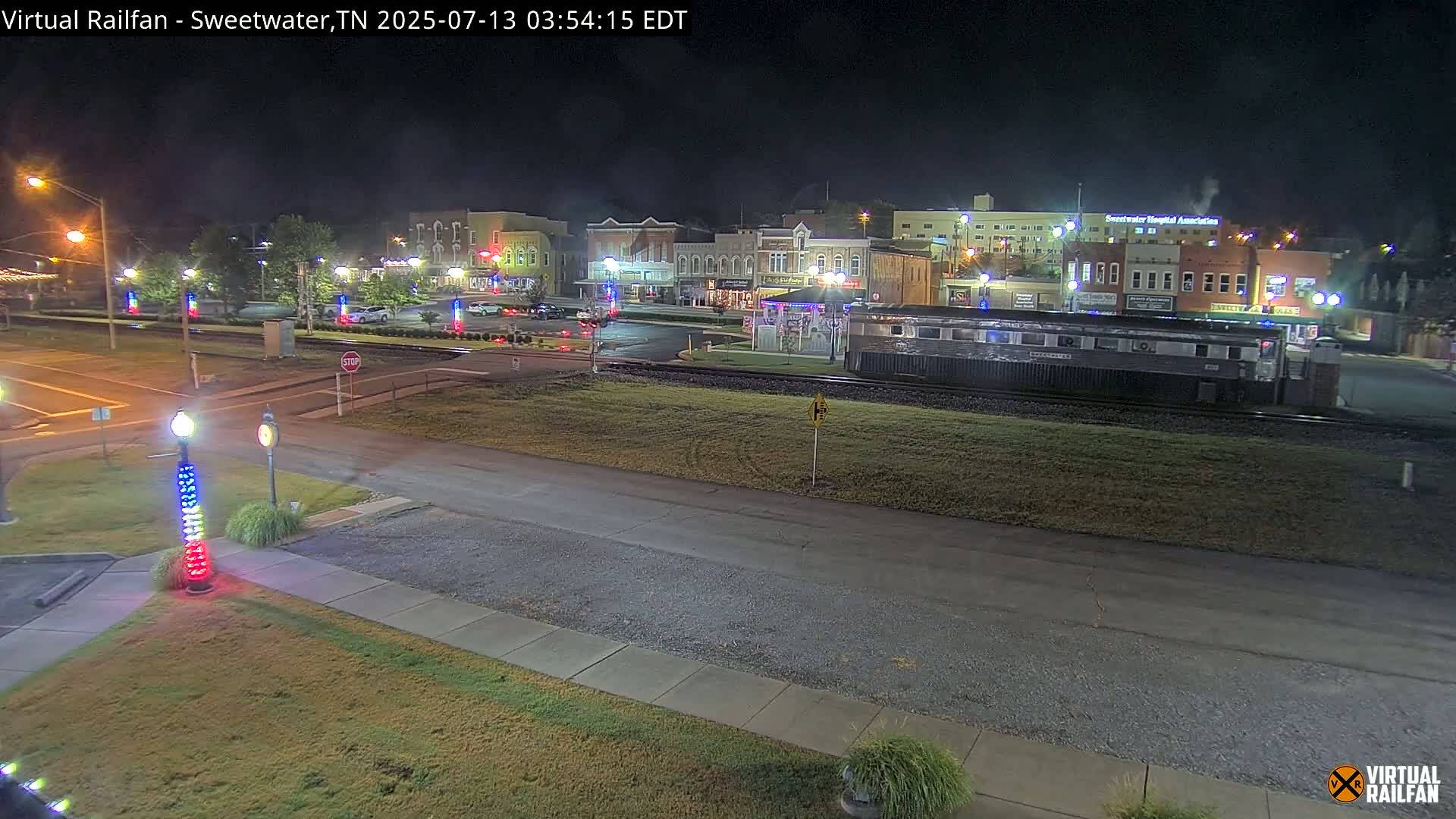 A nighttime view of a town square with several buildings, a railway car, and a few parked cars, under clear skies.