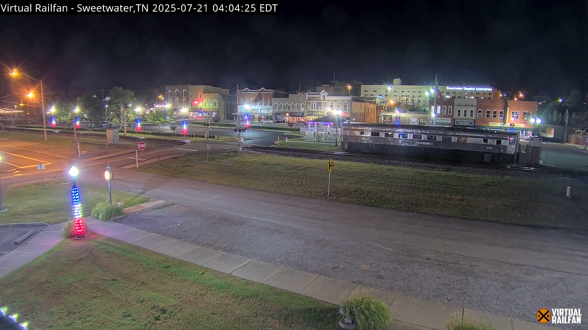 A nighttime view of a town square with several buildings, a train car on tracks, and decorative lights along a roadway.