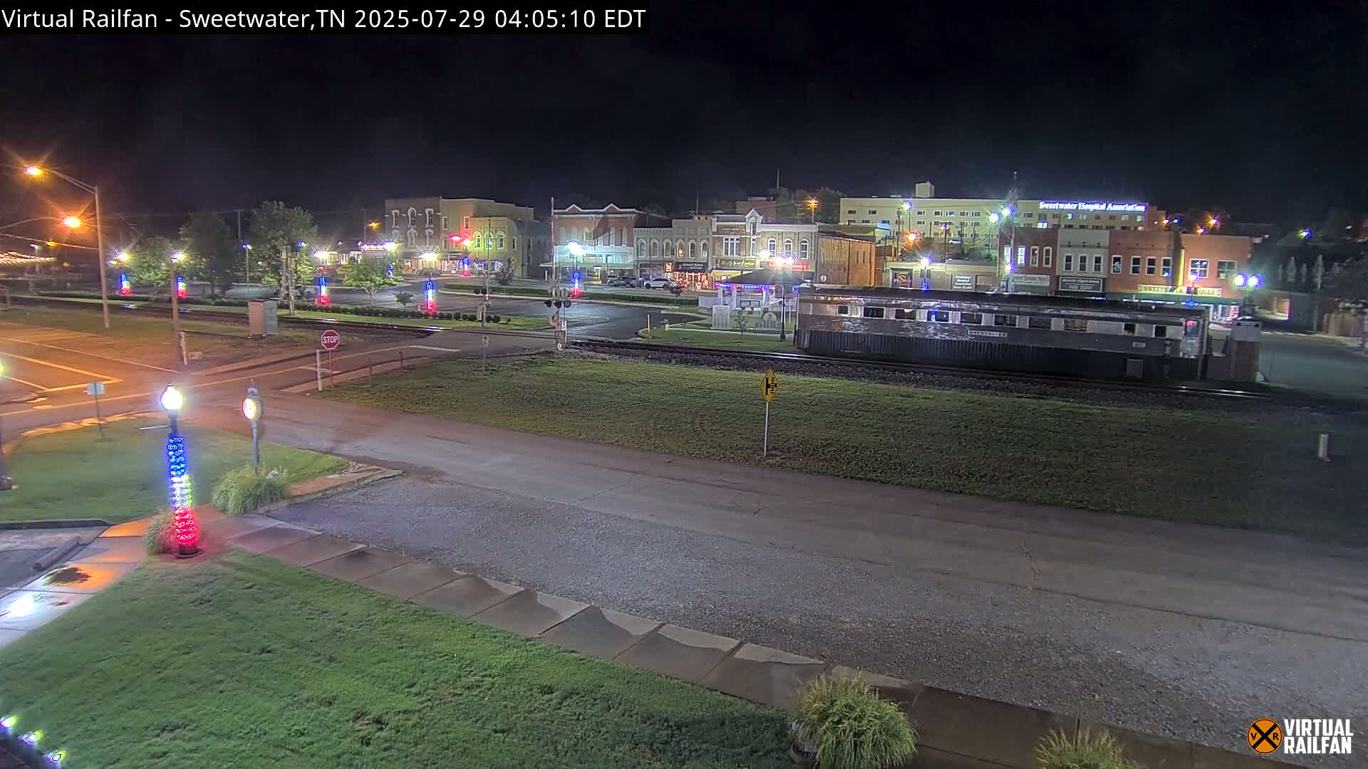 A nighttime view of a town square with a train car parked on the tracks in front of several buildings, illuminated by streetlights.