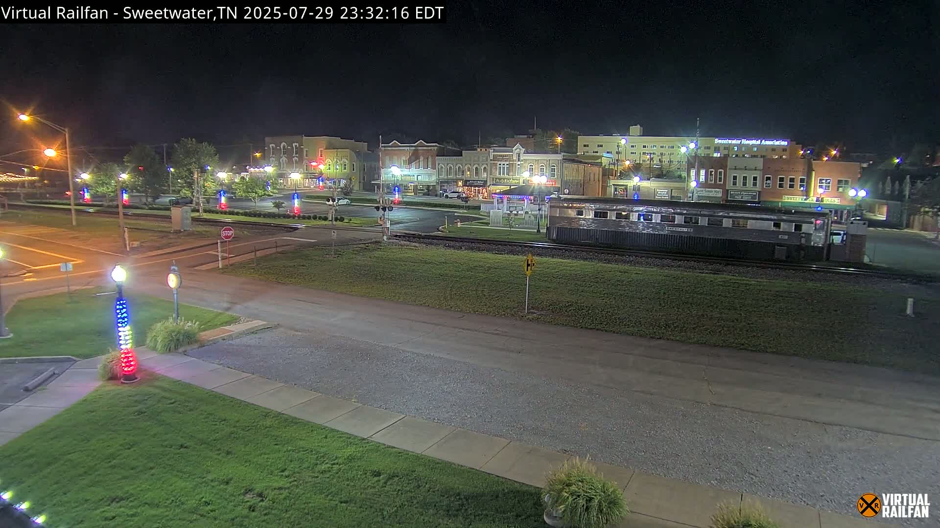 A nighttime view of a town square with a train car parked on the tracks in front of several buildings, illuminated by streetlights.