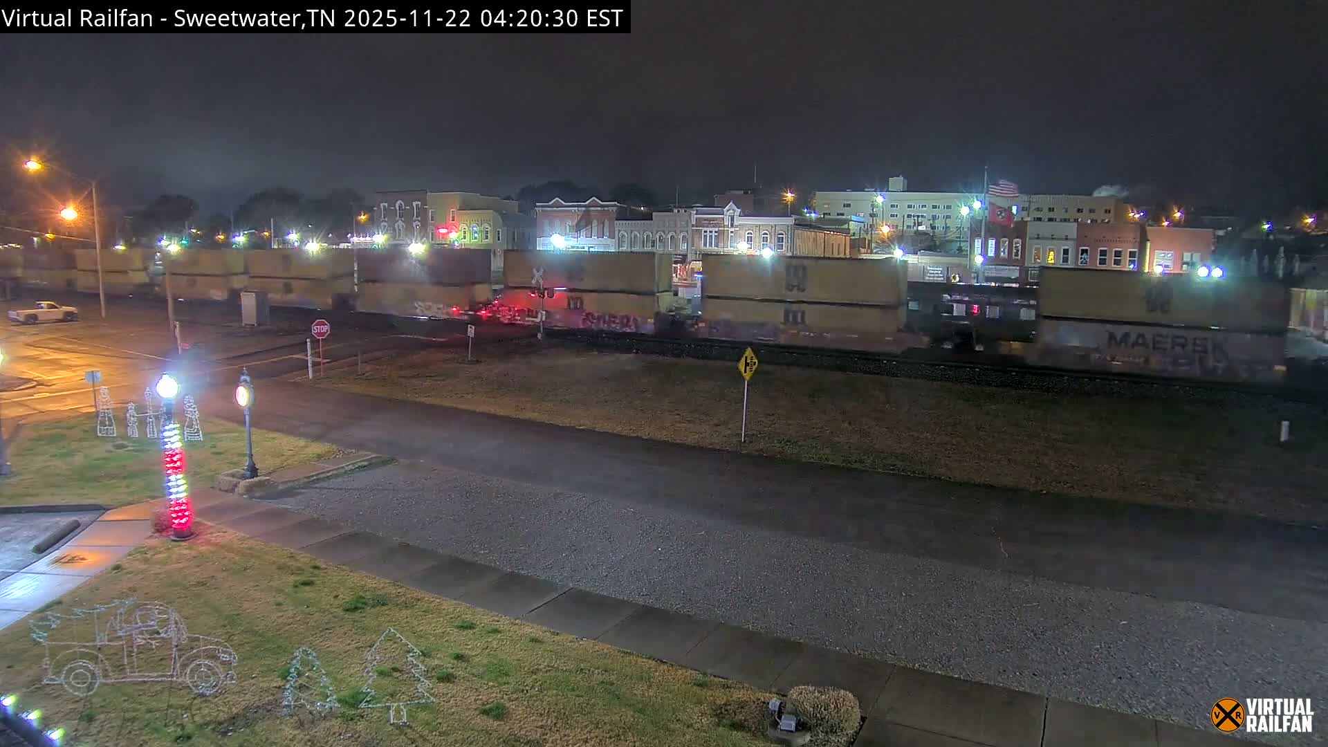 A long train carrying cargo containers travels through a damp, illuminated town at night, past twinkling Christmas light decorations and glowing streetlights.