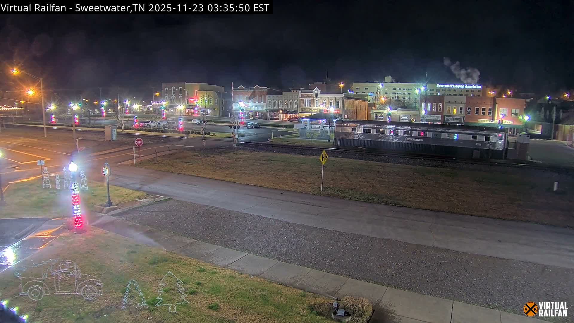 On a clear night, a train rests on tracks alongside a festive small-town street adorned with holiday lights and surrounded by buildings, with a visible plume of steam rising from a distant building.