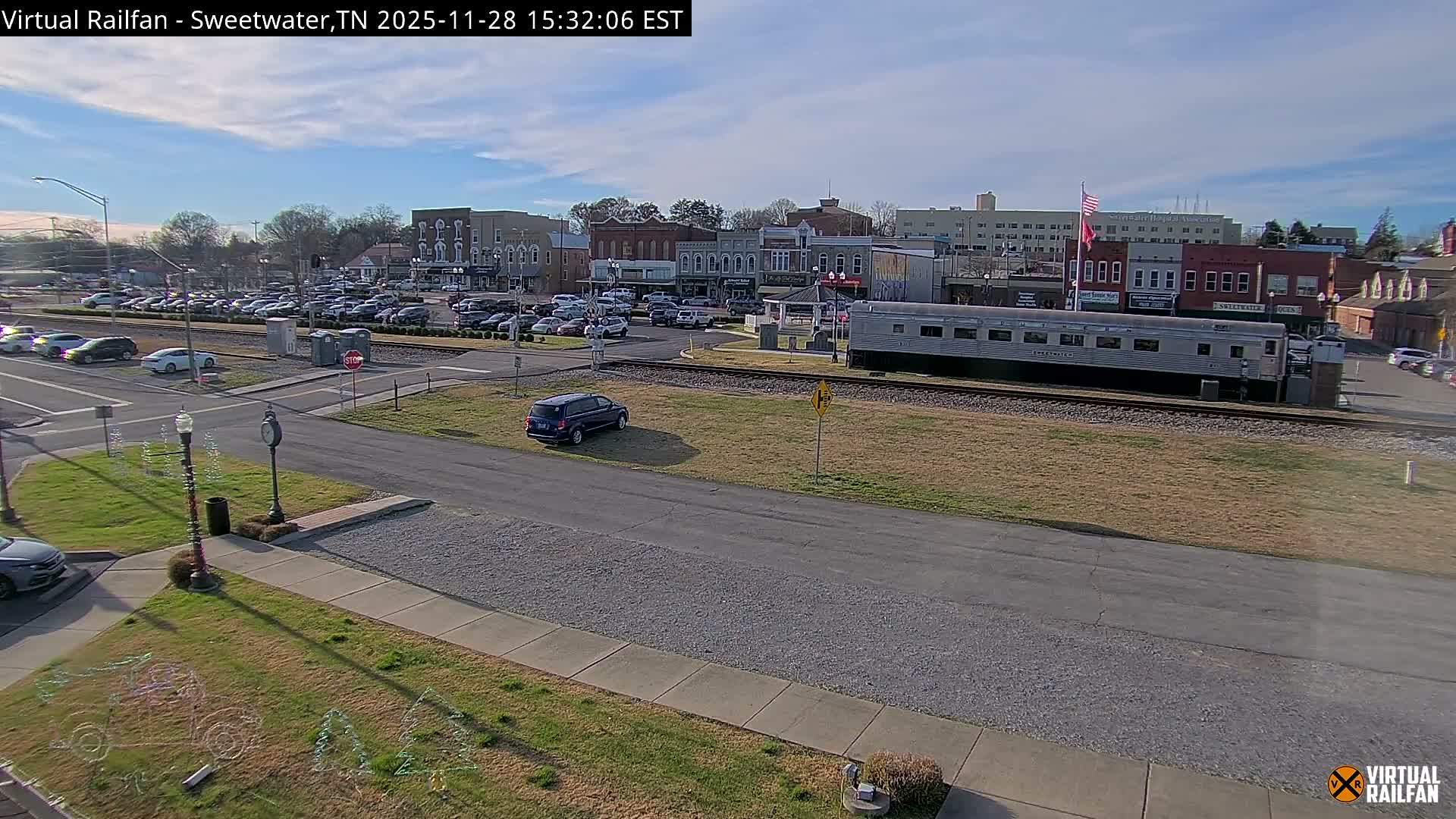 A sunny daytime scene captures a small town featuring a silver train car on tracks, a dark blue SUV parked on a grassy median, a busy parking lot filled with cars, and a backdrop of various buildings under a partly cloudy sky.