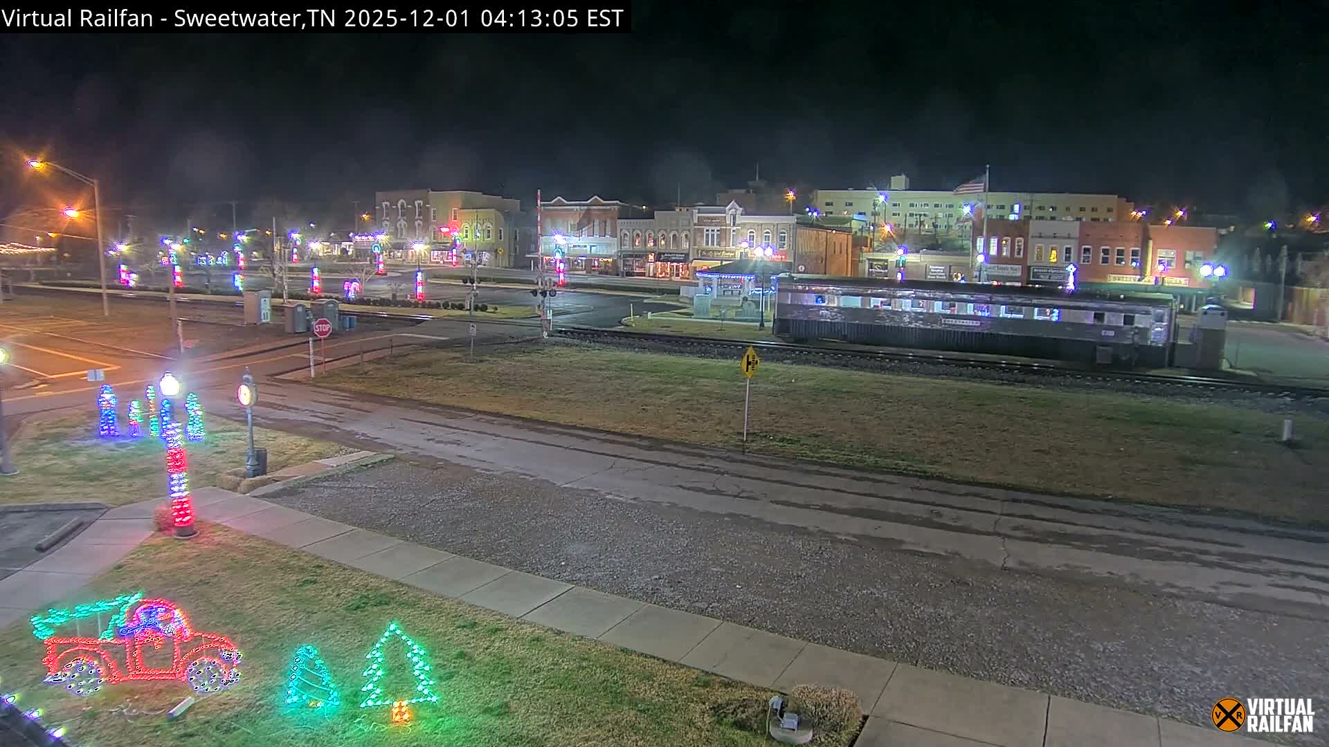 The image displays a clear night view of a festive town decorated with numerous colorful Christmas lights, featuring a train car parked on tracks running through the middle ground, and illuminated buildings lining the street in the background.