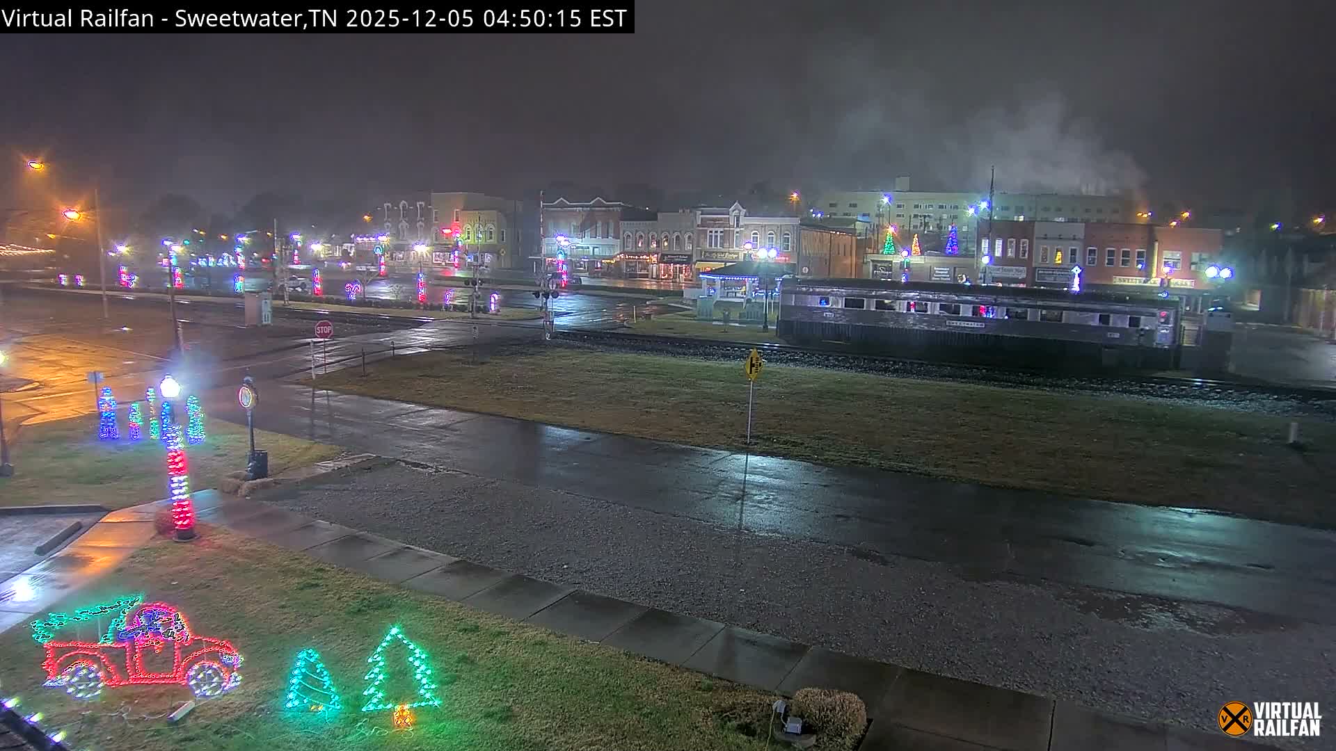 A festive nighttime scene in a town features colorful Christmas lights illuminating wet streets and a train car parked on tracks, with buildings in the background under an overcast and wet sky.