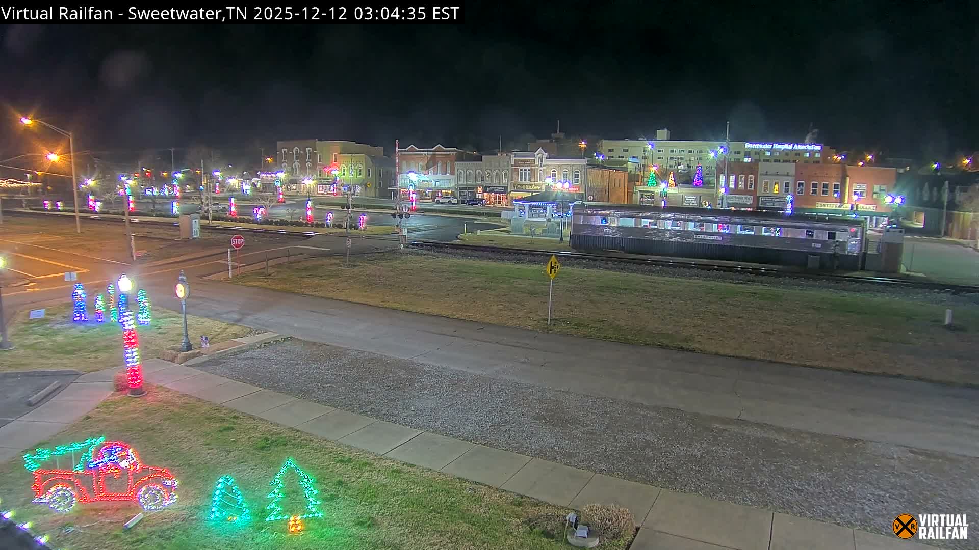 A clear, dark night illuminates a festive small-town scene, where numerous colorful Christmas lights adorn buildings, trees, and ground displays, with a silver train car parked beside the tracks running through the brightly lit area.