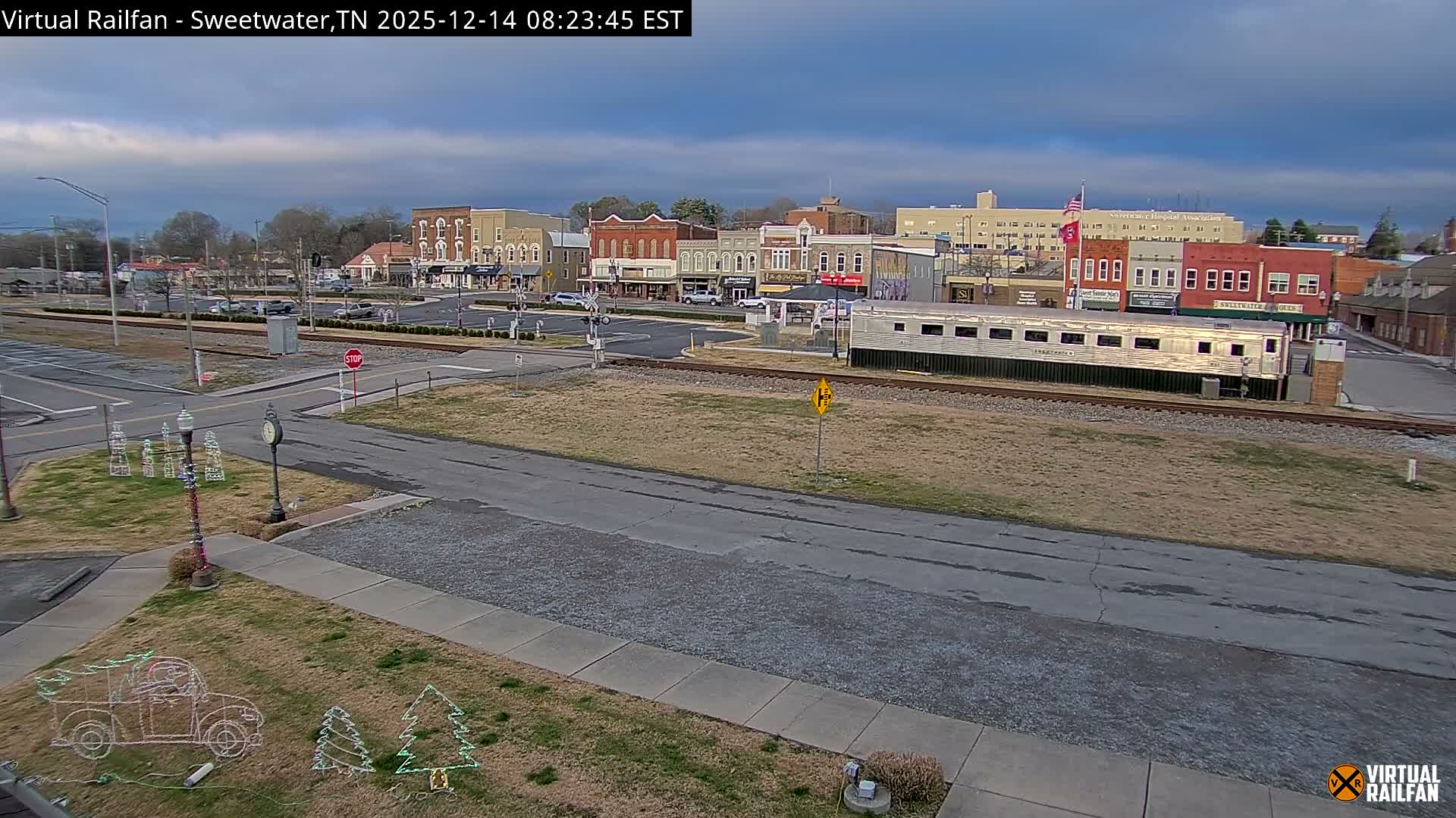A clear, dark night illuminates a festive small-town scene, where numerous colorful Christmas lights adorn buildings, trees, and ground displays, with a silver train car parked beside the tracks running through the brightly lit area.