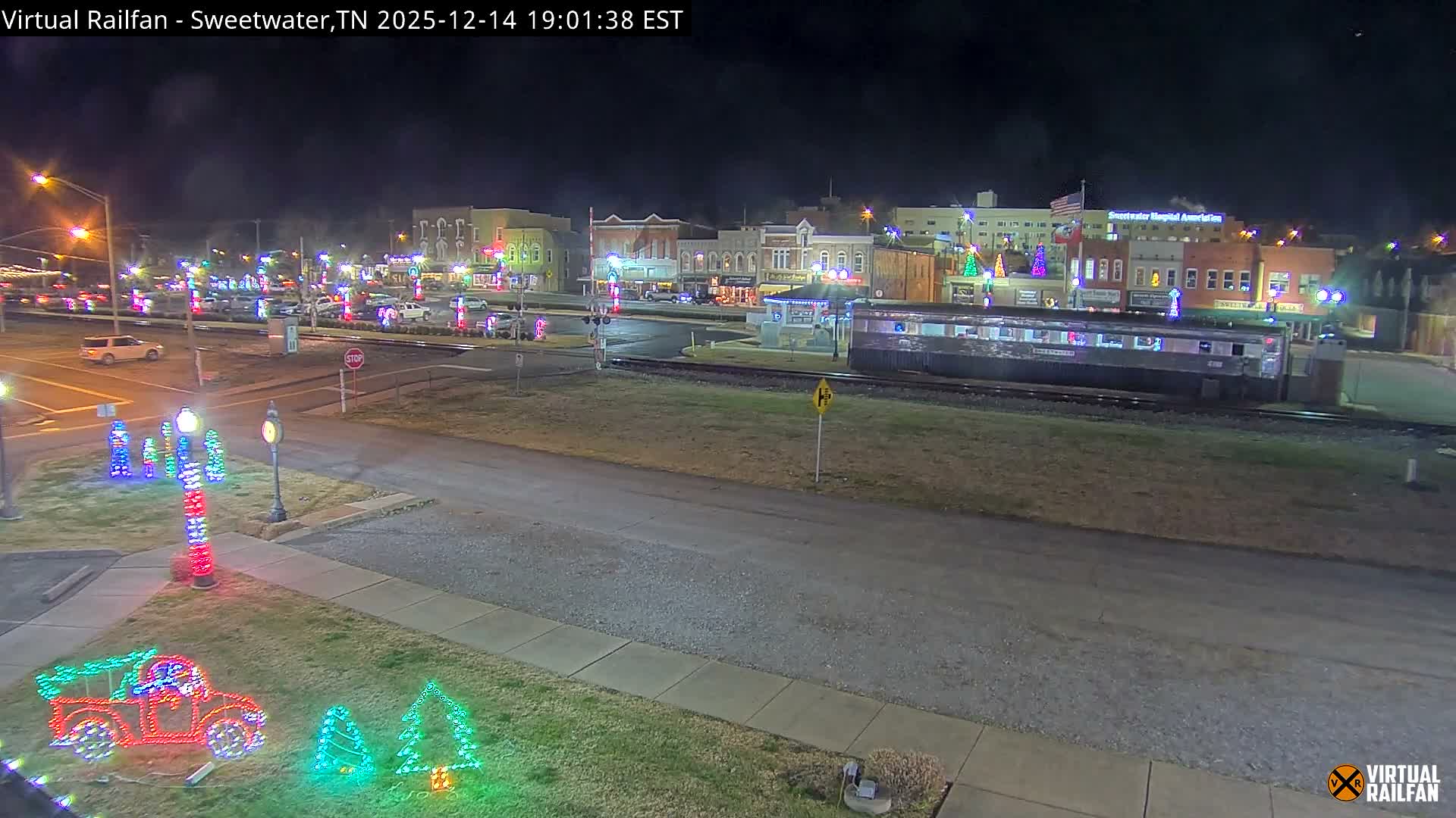 A clear, dark night illuminates a festive small-town scene, where numerous colorful Christmas lights adorn buildings, trees, and ground displays, with a silver train car parked beside the tracks running through the brightly lit area.
