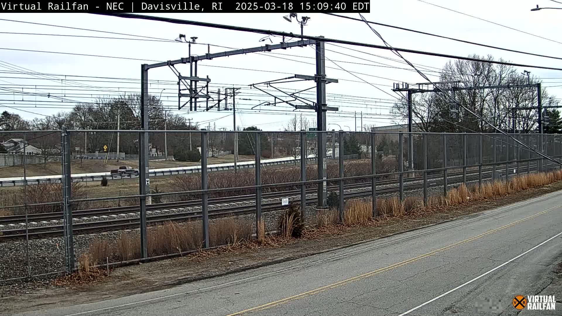 A section of railway tracks runs behind a chain-link fence, alongside a road, under an overcast sky.