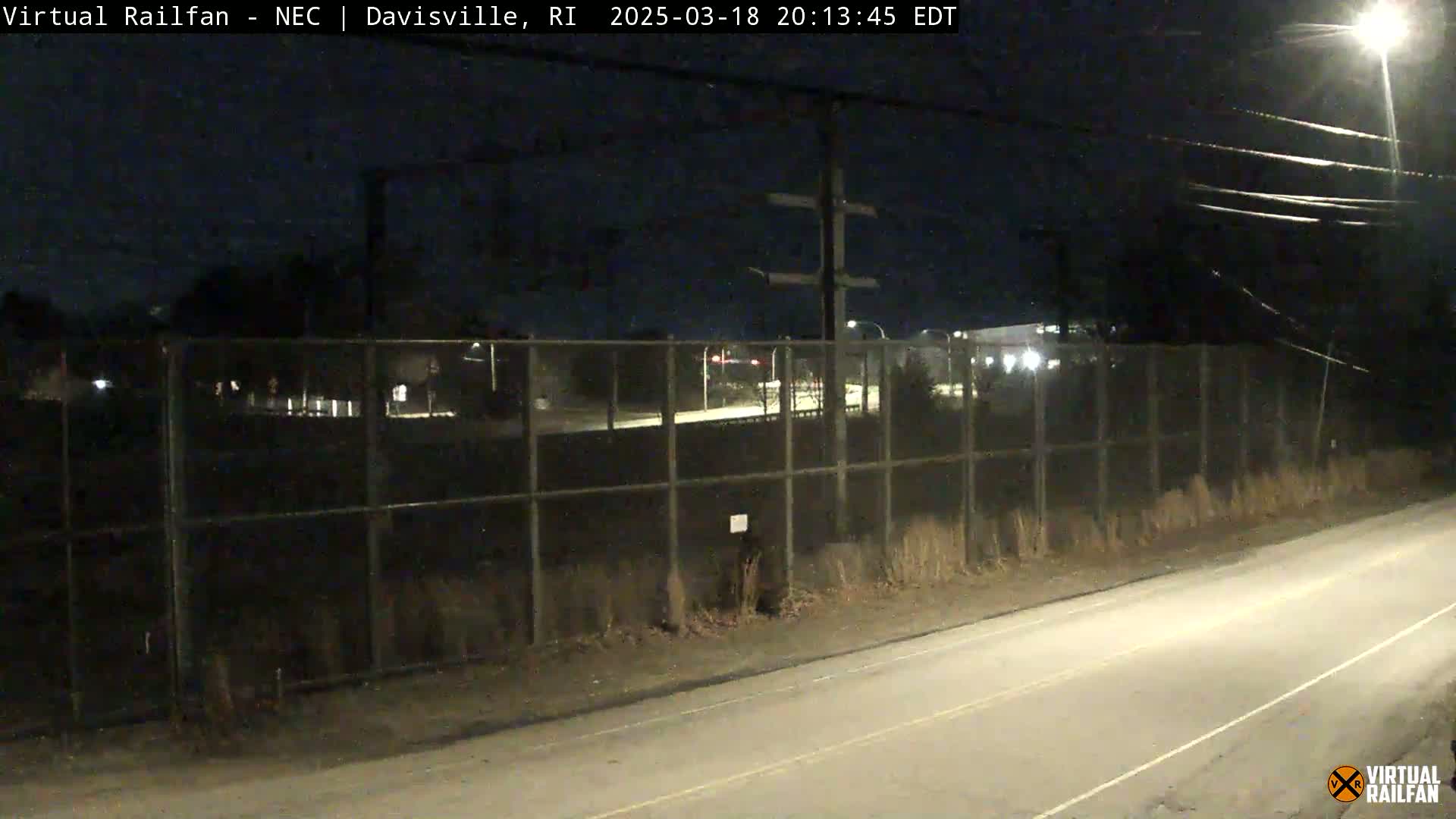 A nighttime view of a road next to a chain-link fence separating it from a dimly lit area beyond, with a clear sky.