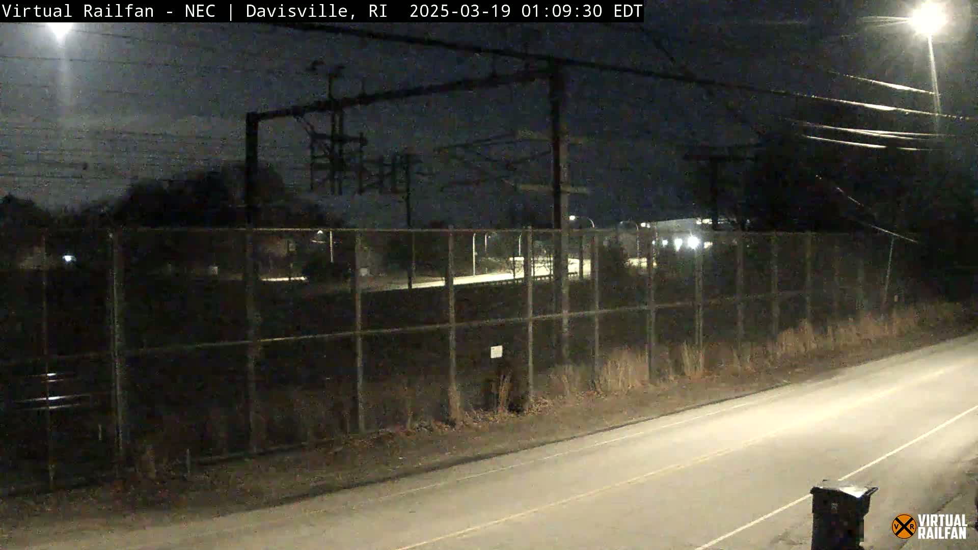 A nighttime view of a chain-link fence separating a road from a railway area under a dark, clear sky.