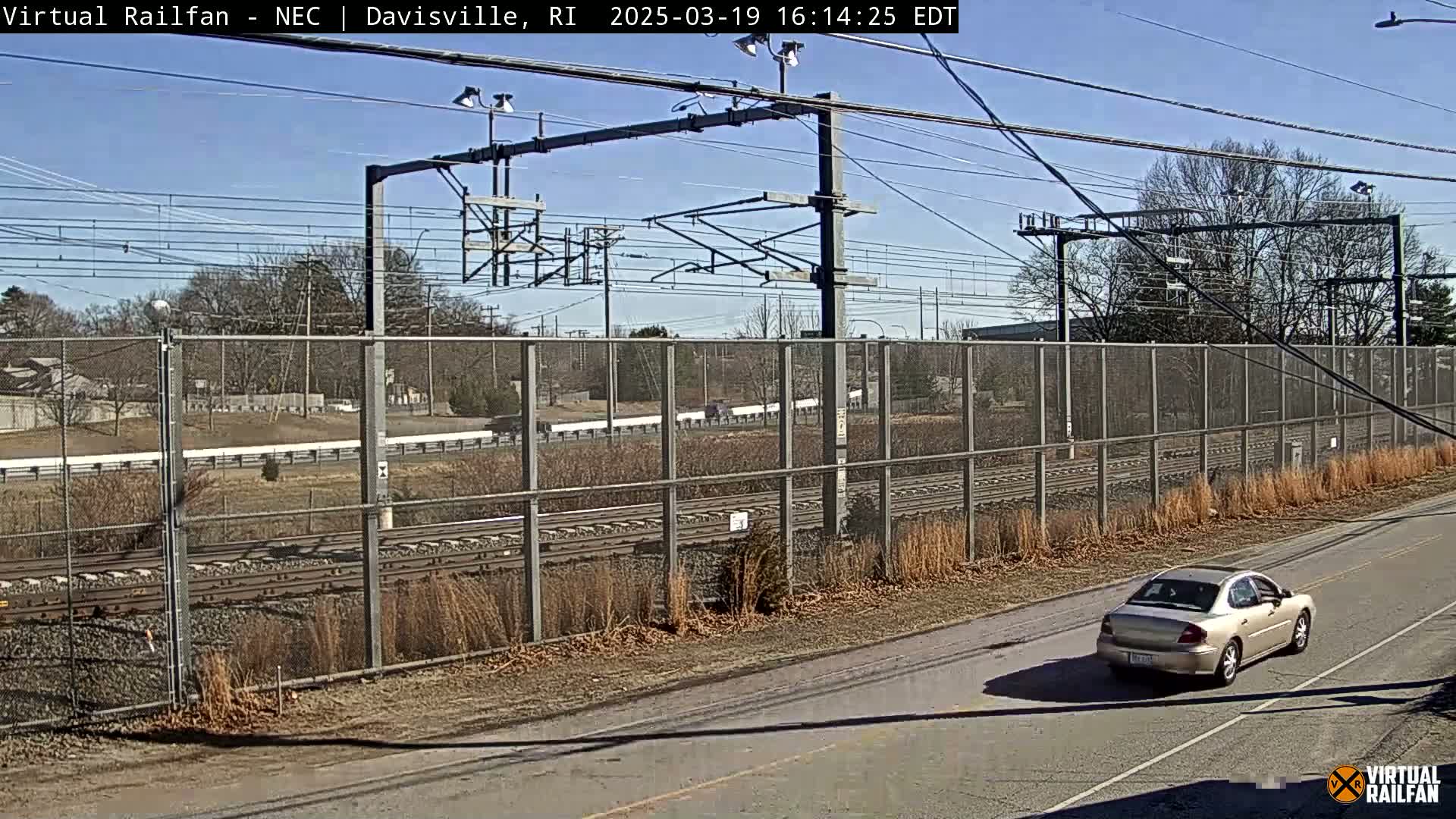 A beige car drives on a road next to a chain-link fence bordering train tracks under a clear, sunny sky.