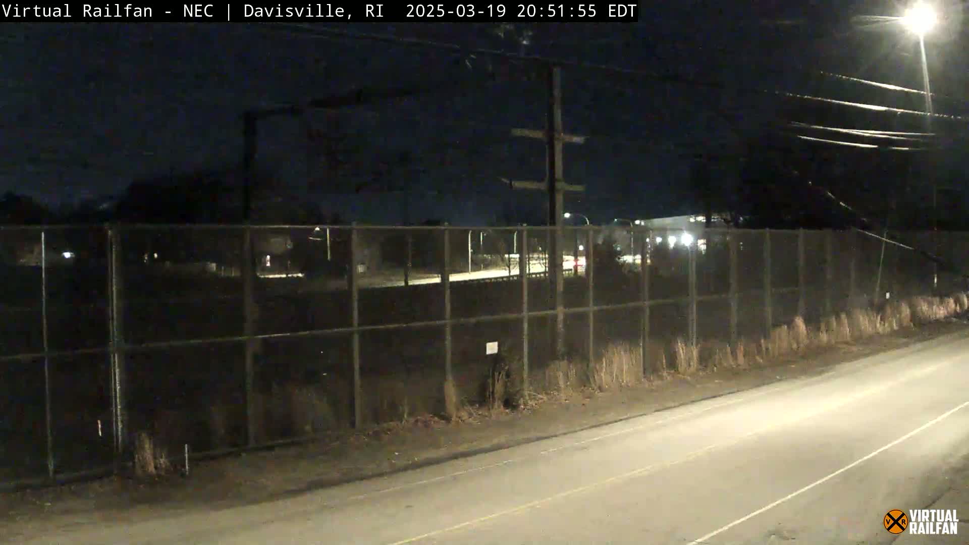 A nighttime view of a road beside a chain-link fence enclosing an area with some lights visible beyond it.
