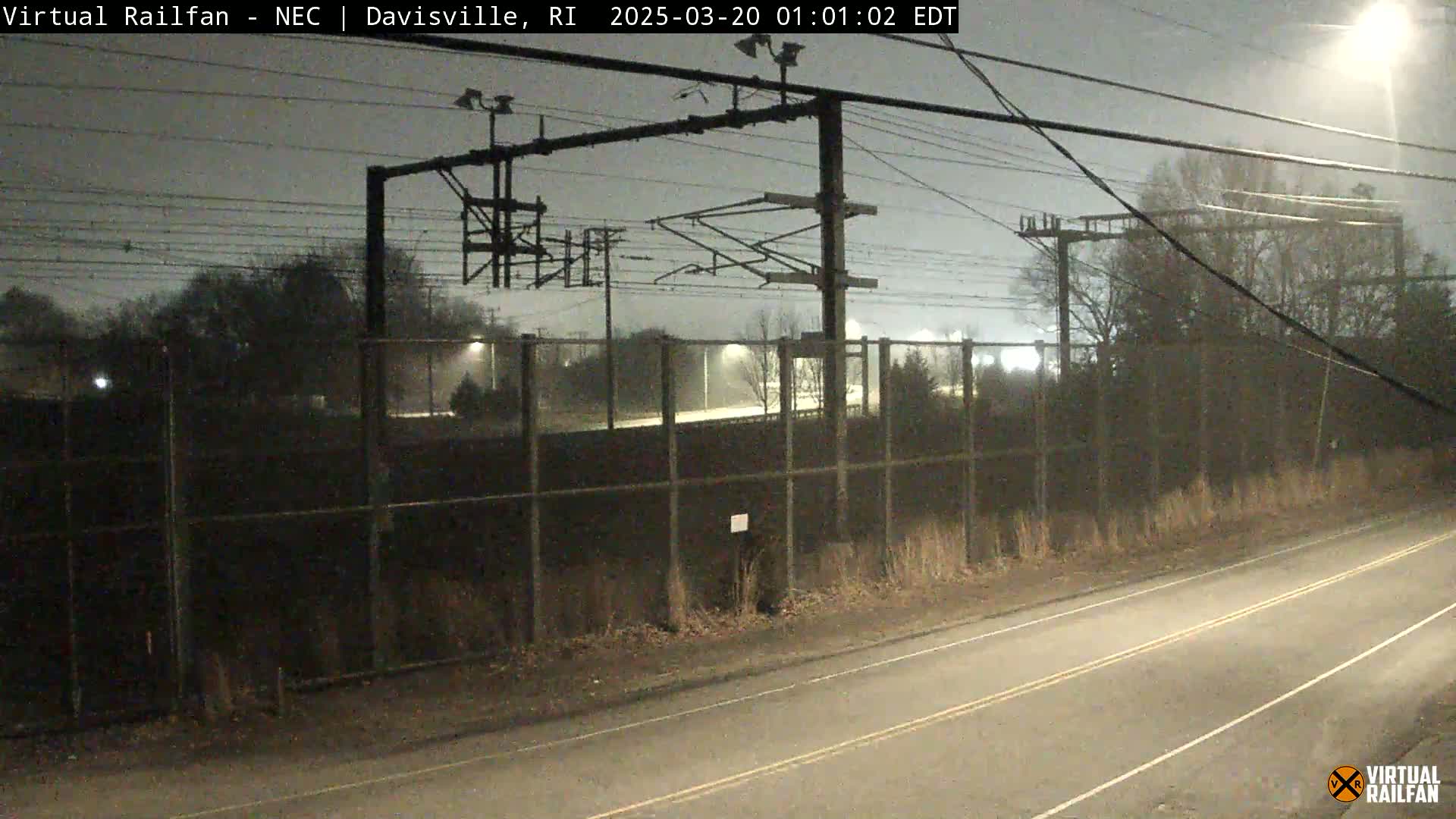 A nighttime view of a road next to a chain-link fence enclosing a railway area with overhead power lines, under overcast skies.