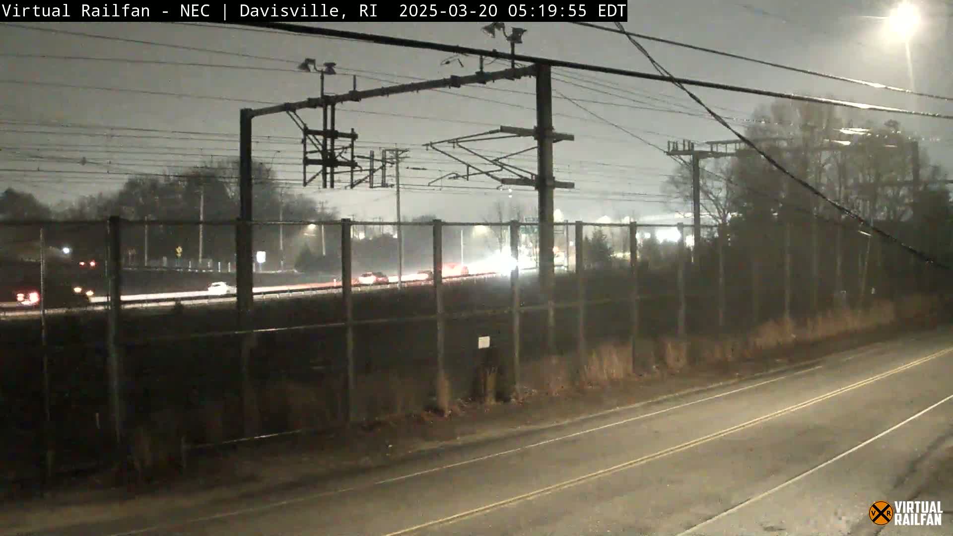 A nighttime view of a highway with sparse traffic behind a chain-link fence, under a dark, overcast sky with light precipitation.