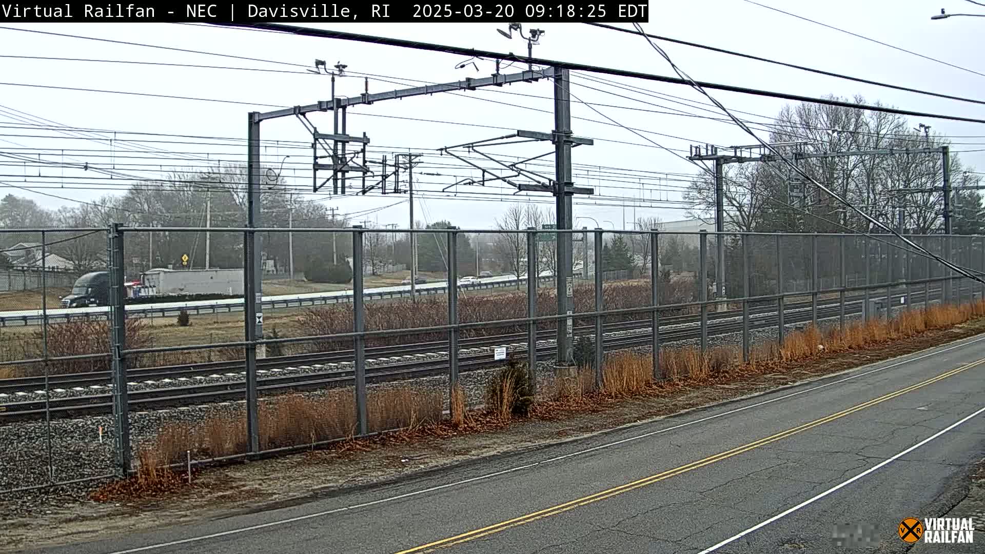 A view of a road beside train tracks on an overcast day, with a chain link fence separating them, and sparse, dry vegetation along the roadside.