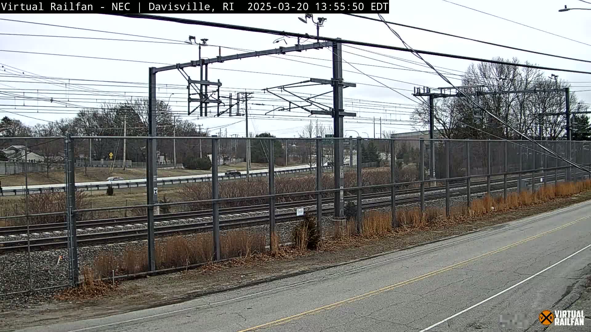A view of a railway line behind a chain-link fence next to a road, on an overcast day.