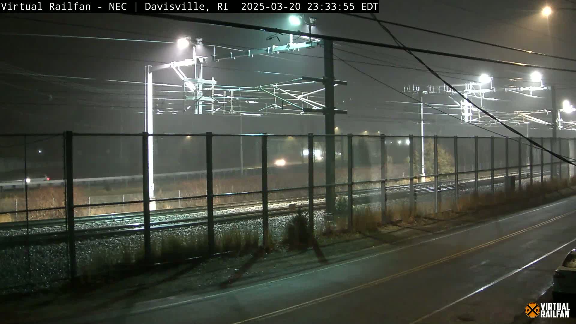 A nighttime view of a train track behind a fence, illuminated by lights in foggy conditions.