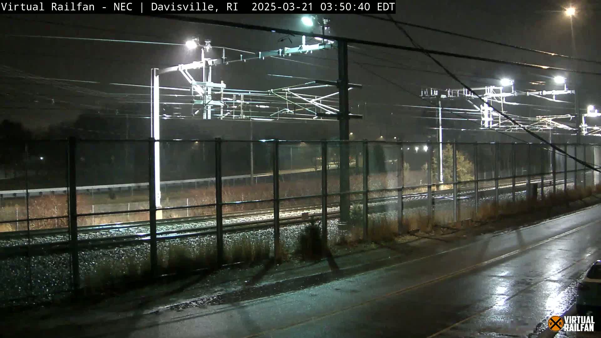 A nighttime view of rain-slicked train tracks behind a chain-link fence, illuminated by overhead lights.