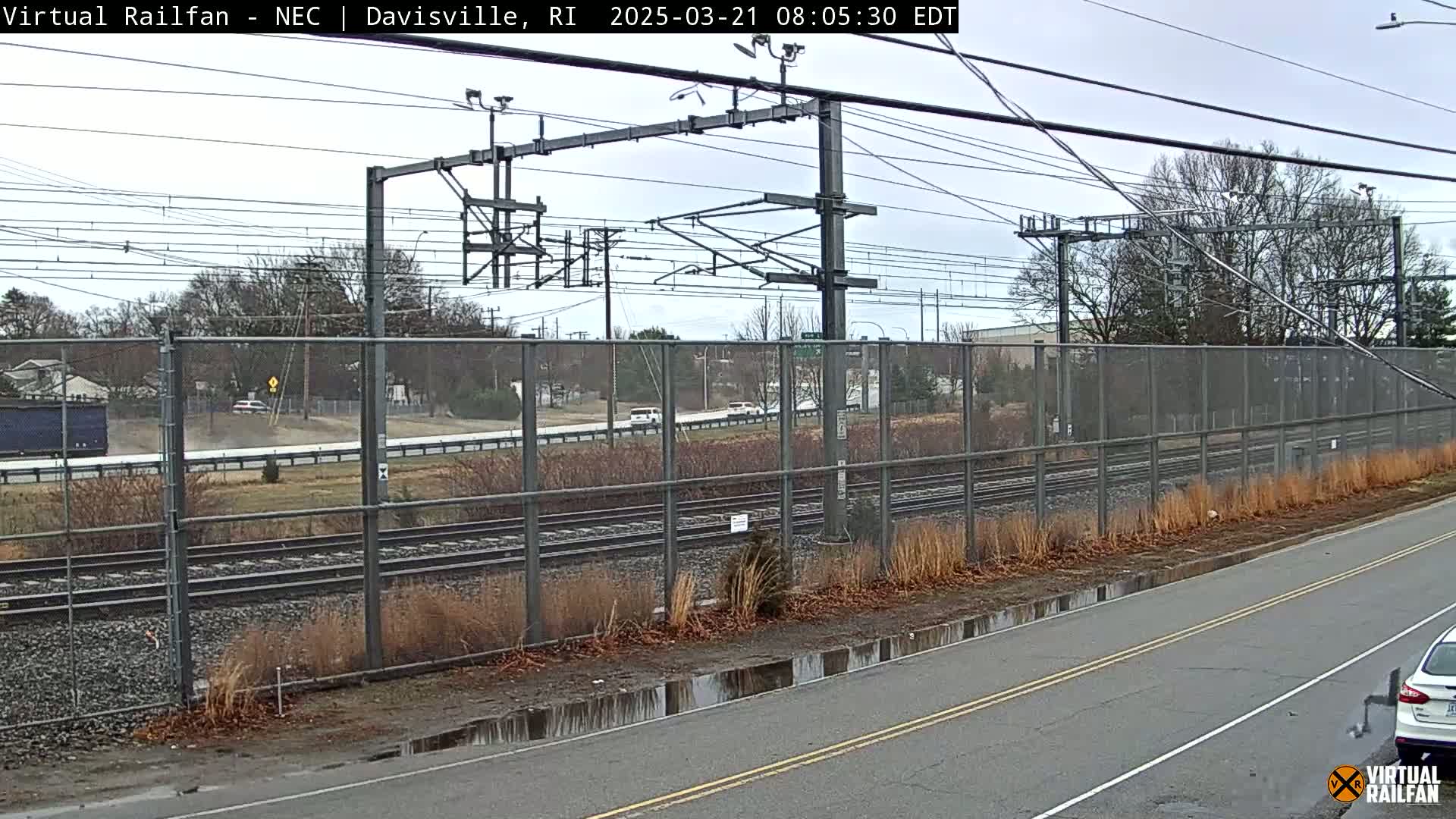 A view of a railroad track behind a chain-link fence on an overcast day with puddles on the road.