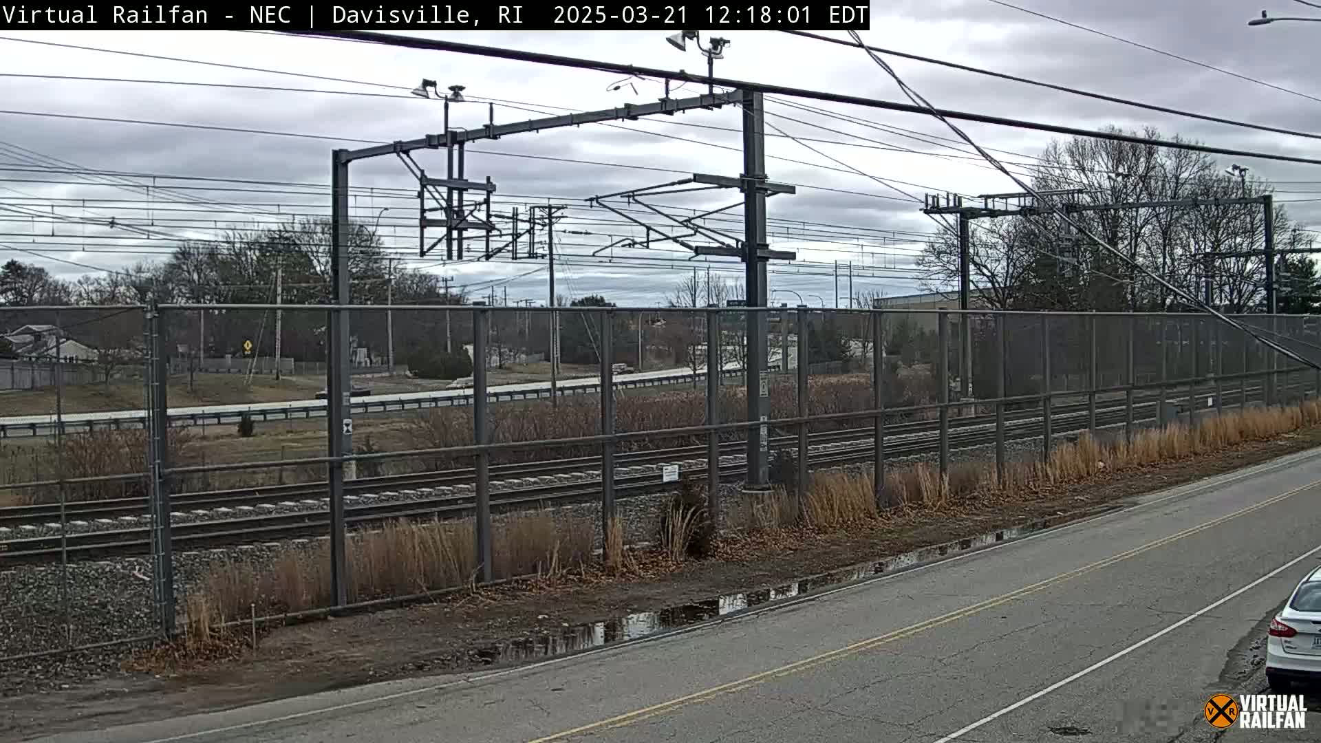 A view of railroad tracks behind a chain link fence on an overcast day, with puddles on a nearby road.