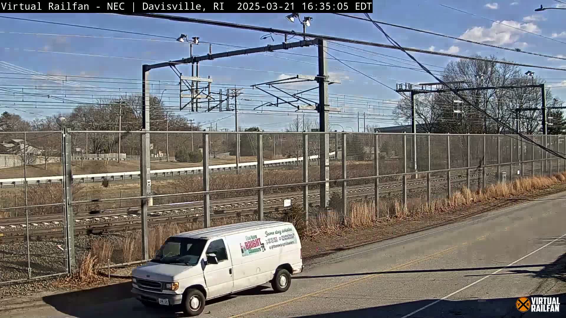 A white van is parked on a road next to a chain-link fence bordering railroad tracks under a sunny sky.