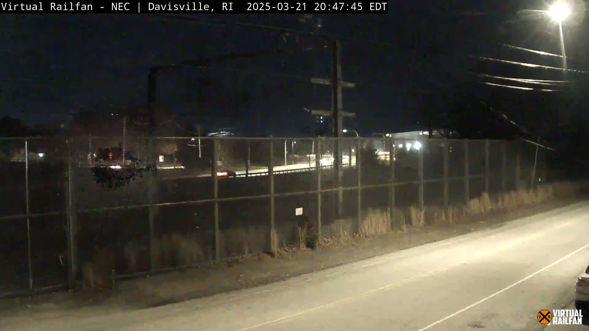 A nighttime view of a road and a chain-link fence separating it from a roadway beyond, with sparse vegetation and a few vehicles visible under dark, clear skies.