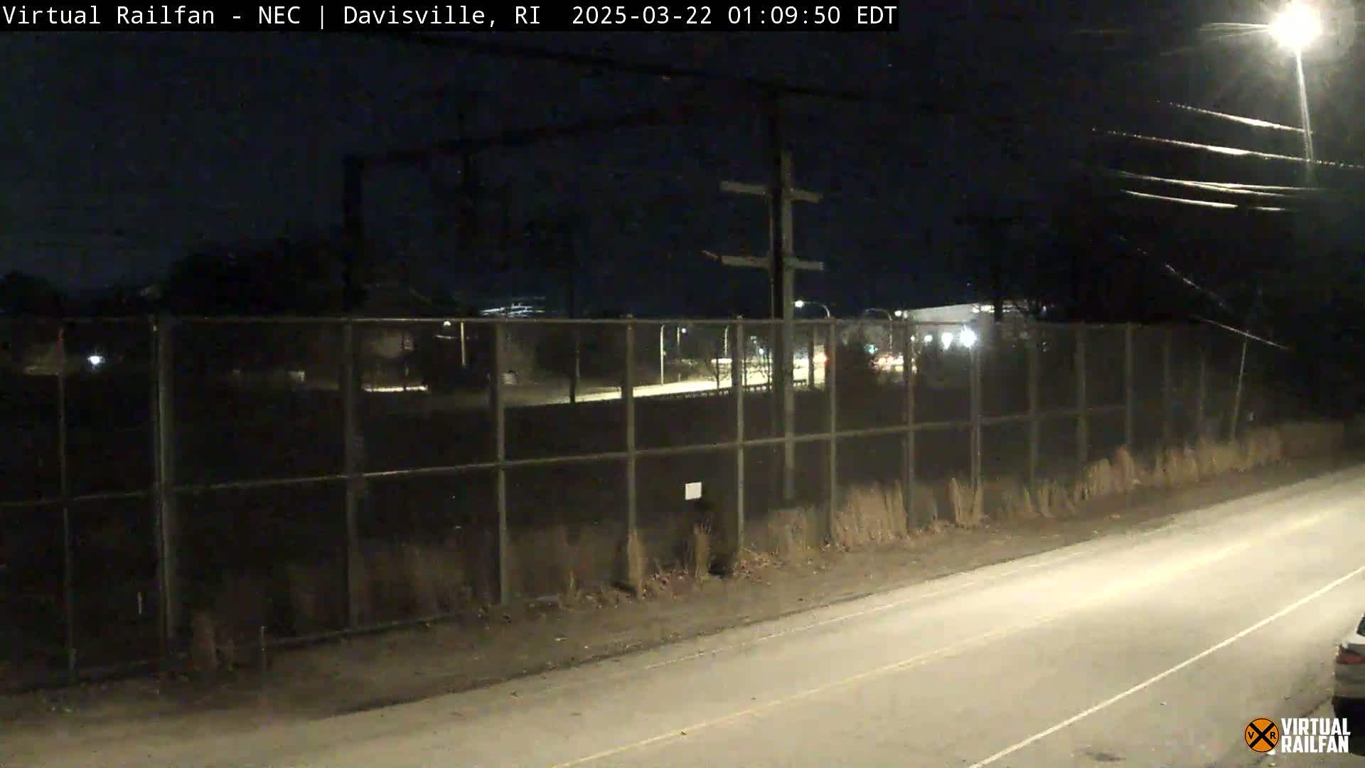 A nighttime view of a road next to a chain-link fence, beyond which are dimly lit buildings and power lines.